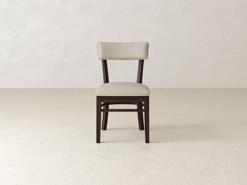 A single wooden chair with a cushioned back and seat is centered against a plain, light-colored wall and floor. The chair has dark legs and a light beige cushion.