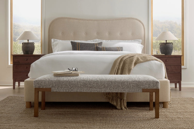 A bedroom features a large James & James Florence Bed in beige velvet, white bedding, and a plush gray textured bench. Wooden nightstands with lamps flank the bed, two large windows provide natural light, and a woven rug enhances the room's modern appeal.