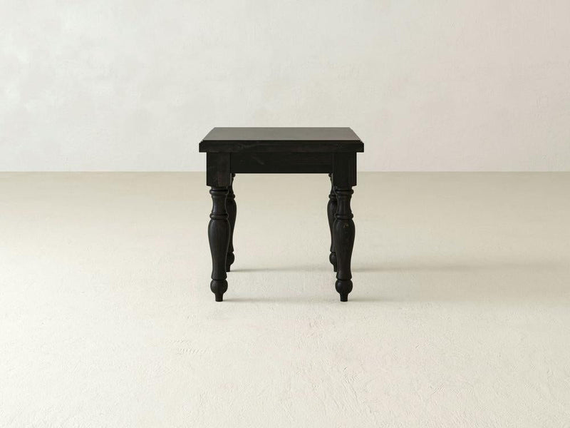 The Abigail End Table - Charred Ember by James+James, featuring ornate, turned legs in a black finish, stands on a plain, light-colored floor against a white wall. The table boasts a classic, vintage design with a smooth, rectangular surface.
