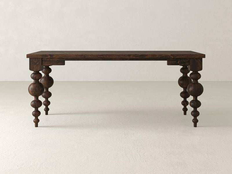 The Olivia Desk - Tobacco by James+James, featuring dark wooden construction and intricately carved legs with spherical and cylindrical shapes, is set against a plain, light-colored background. Its simple rectangular top combined with the detailed craftsmanship on the legs gives it an elegant antique appearance.