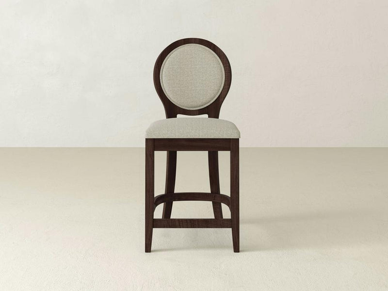 The Alice Stool by James & James offers modern elegance with its light gray circular backrest and plush seat. It features a sleek maple wood frame, four straight legs, and a simple footrest, all displayed against an off-white background.