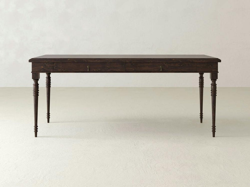 The Audrey Desk from James+James is a dark wooden table featuring four intricately carved legs, set against a plain, light-colored background. Its rectangular surface and two subtle front drawers enhance its timeless elegance, making it a classic addition to any space.
