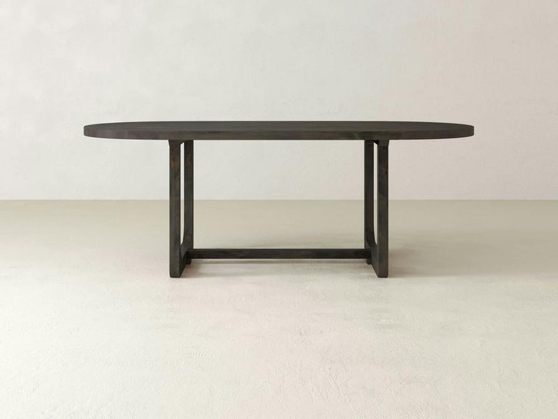 The James+James Emersen Oval Dining Table is crafted from knotty alder hardwood, showcasing a simplistic design with modern elegance. Its sturdy base features two vertical supports connected by a horizontal bar. The background highlights the table with light, neutral-colored walls and floors.