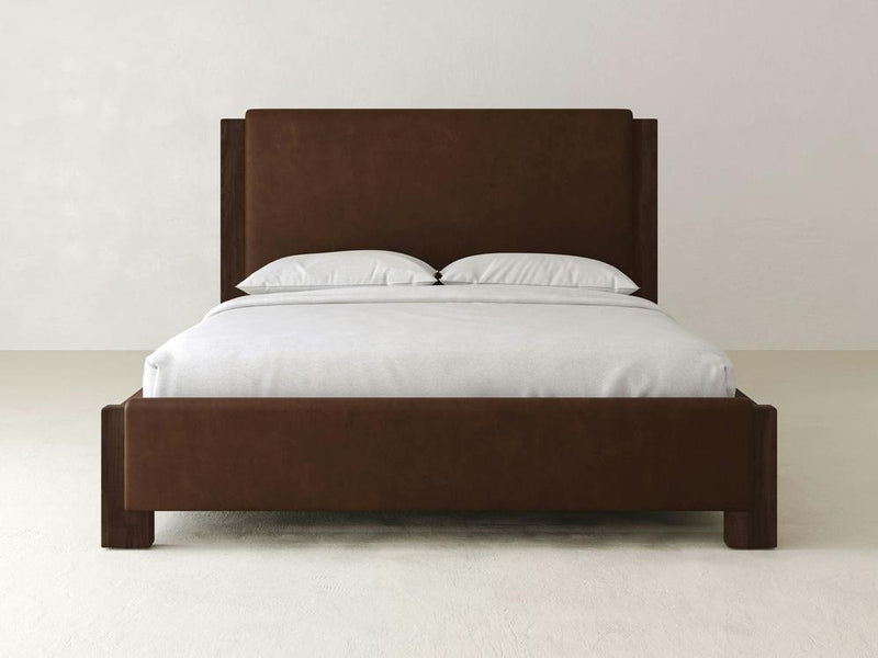The Lennon Bed – Leather by James & James is a modern piece featuring durable leather and a brown wooden frame. Its tall headboard and low footboard pair elegantly with white pillows and a light gray comforter, against a plain, light-colored wall.