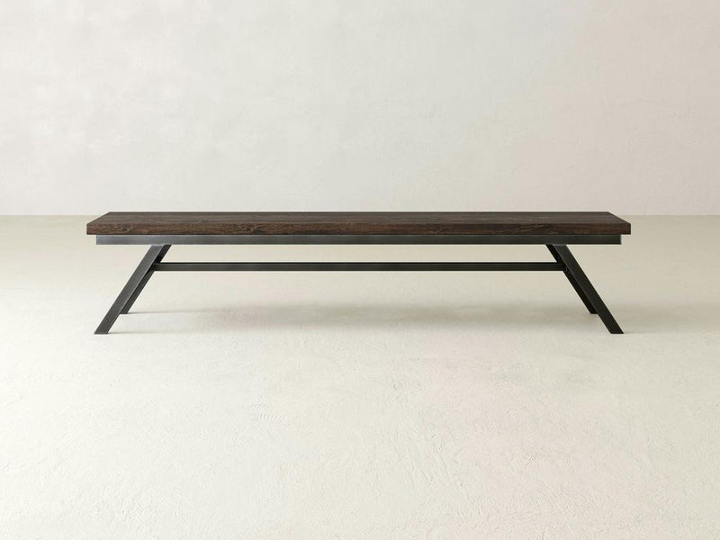The Fulton Bench - Tobacco by James+James, featuring a modern and minimalist design with a dark brown wooden top and black angular metal legs, is placed against a plain white background. This sleek bench boasts a clean design that's perfect for contemporary settings.