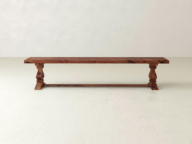 Against a plain beige wall stands the Heirloom Bench - Tuscany from James+James. This wooden bench showcases a rectangular seat with a smooth finish, supported by two intricately carved legs connected by a horizontal bar at the base. The overall design combines minimalist and rustic elements.