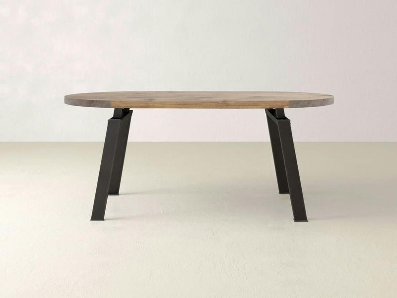 The Fulton Oval Dining Table by James+James features a modern industrial design with an oval, solid hardwood top and a smooth light brown surface. It is supported by four angled raw steel legs, creating a minimalist and contemporary look, all set against a neutral, light-colored background.