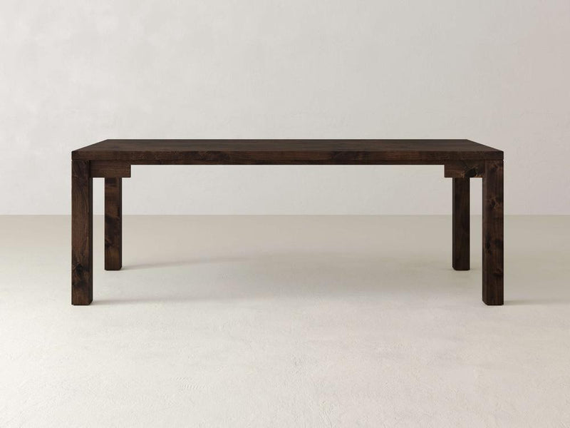 The Jonathan Desk by James+James is a rectangular farmhouse-style desk with a dark finish, crafted from solid hardwood. It features four sturdy legs and a minimalist design, resting on a plain light-colored floor against a white wall.