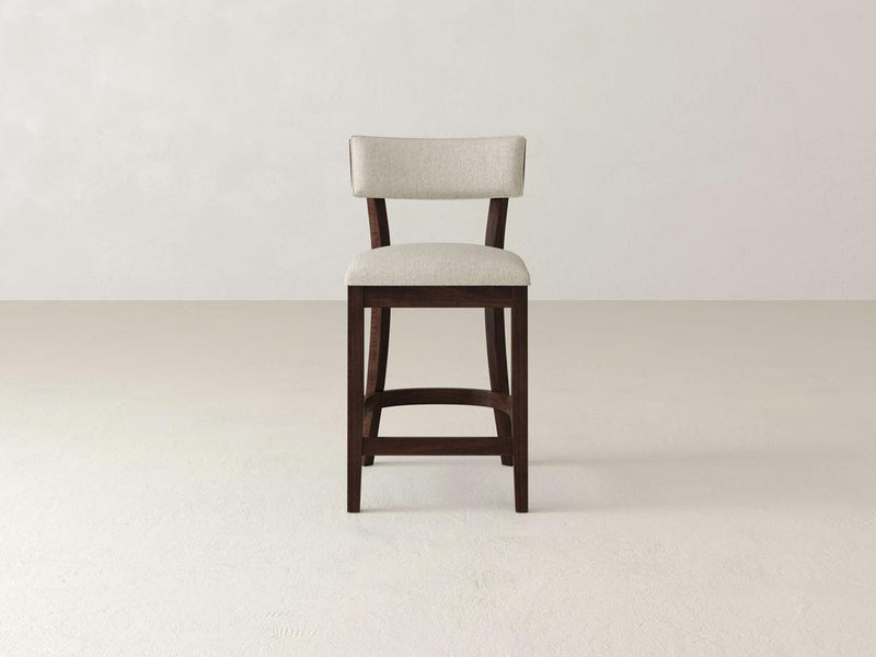 The Everly Upholstered Stool by James & James, with its mid-century design and beige cushioned backrest, showcases minimalist elegance with a rich dark finish. It stands on a light-colored floor against an off-white textured wall.