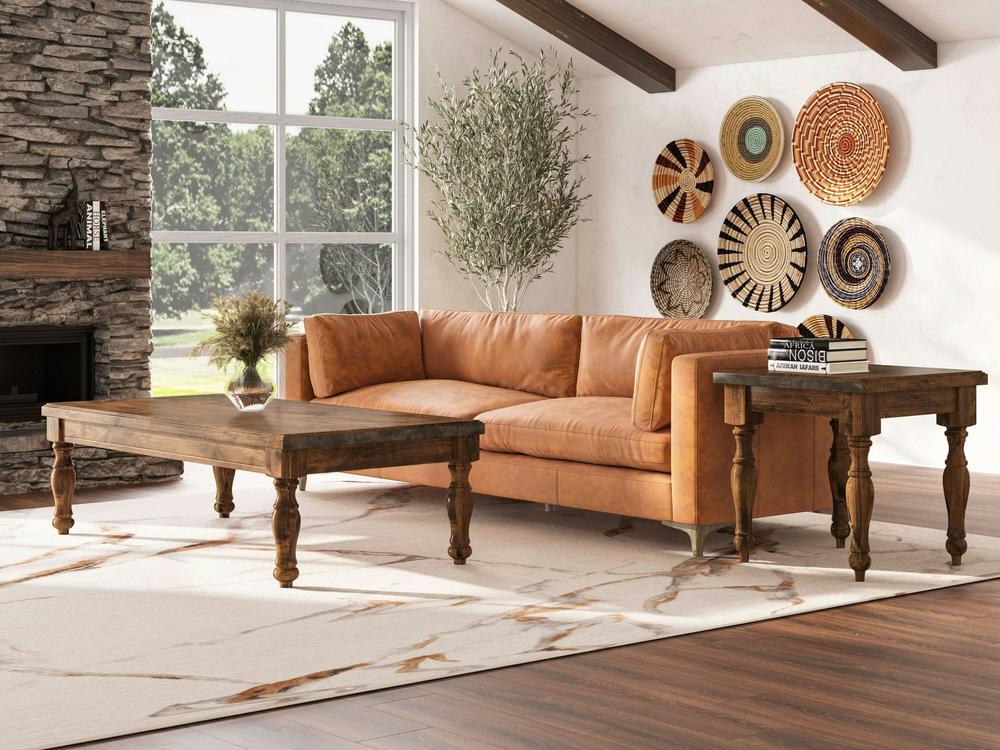 A stylish living room featuring a tan leather sofa, the James+James Abigail Coffee Table in Tuscany, and a side table. The space is accented with a large window, a stone fireplace, a potted plant, and a collection of woven baskets displayed on the wall. The floor has a marble-patterned rug.
