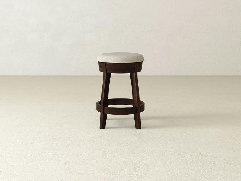 A wooden Jasper Swivel Stool from James & James, with a beige upholstered seat and circular footrests, sits in an empty room with light textured walls and floor. Its swivel function enhances its simple, elegant design.