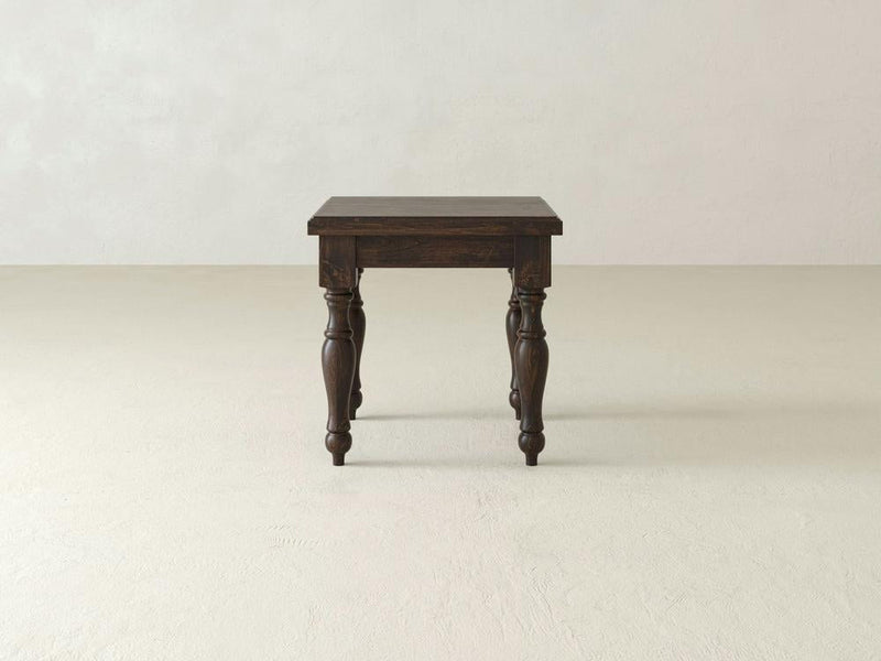 The Abigail End Table - Tobacco by James+James stands on a plain, light-colored, textured floor. The table features a rectangular top and four ornately carved legs. The background is a simple, light-colored wall.