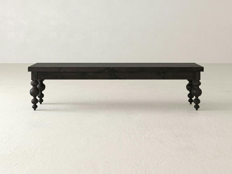 The Olivia Bench - Charred Ember by James+James features a rectangular seat and four intricately carved, spindle-style legs. The bench, finished in a smooth and polished black, boasts a simple yet elegant design that is perfect for indoor use. It stands out beautifully against the plain white background.