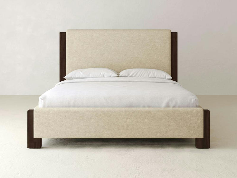 The Lennon Bed – Linen by James & James features a minimalist design with a beige upholstered headboard and dark wooden frame, highlighted by subtle maple accents. It sits in a simple, light-colored room adorned with crisp white linens and two plush pillows.