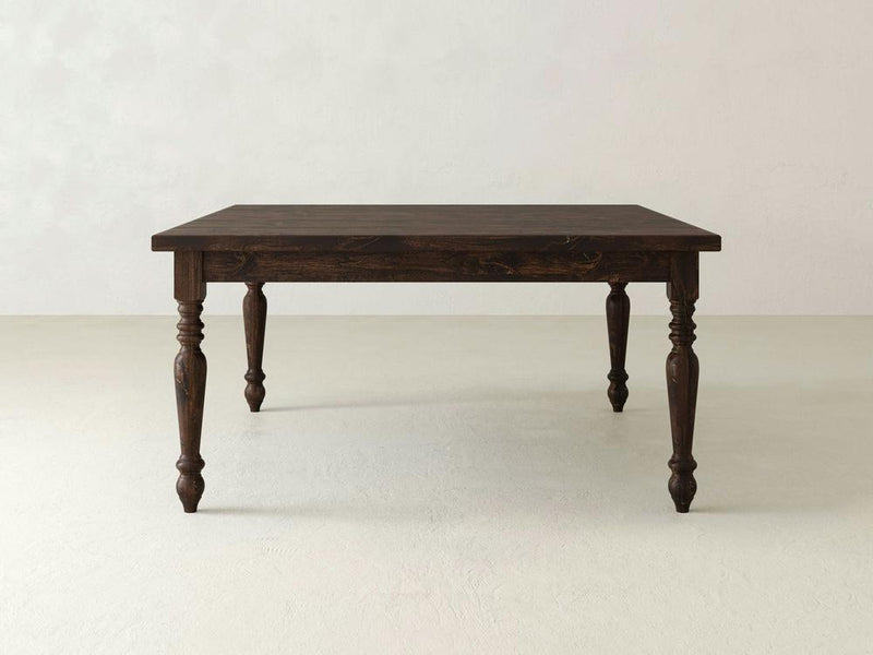 A French Country Square Dining Table in Tobacco finish from James+James stands on a light-colored floor against a plain, light-colored wall. The table features ornate, carved legs and a square top with a classic design, giving it a vintage or rustic appearance.