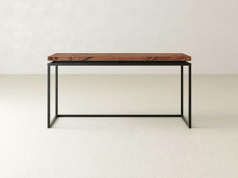 The Floating Top Sofa Table - Tuscany by James+James, featuring a minimalist design with a rectangular wooden top and a black metal frame, stands against a plain off-white wall on a light-colored floor. The table boasts a simple, modern aesthetic with no additional features or decorations.
