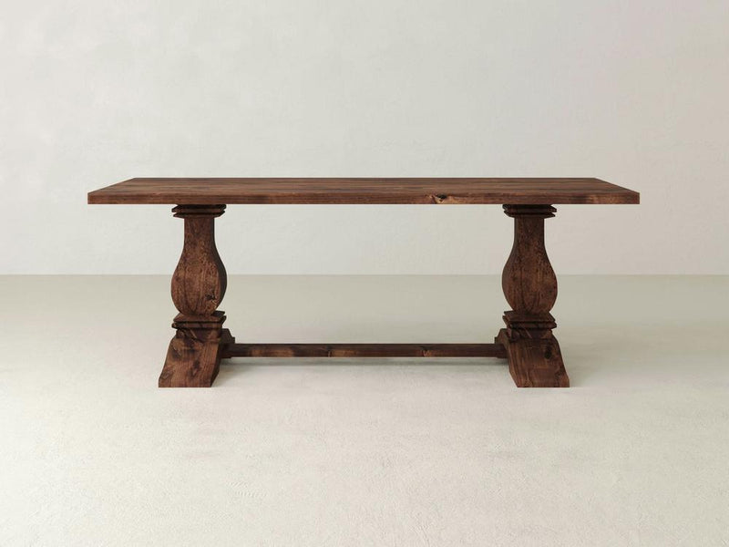 The Heirloom Dining Table - Tuscany by James+James features a rustic wooden design with a flat rectangular top and two ornate pedestal legs connected by a horizontal support. This table showcases a natural wood grain finish and is perfectly set on a light, neutral tiled floor against a plain, light-colored wall.