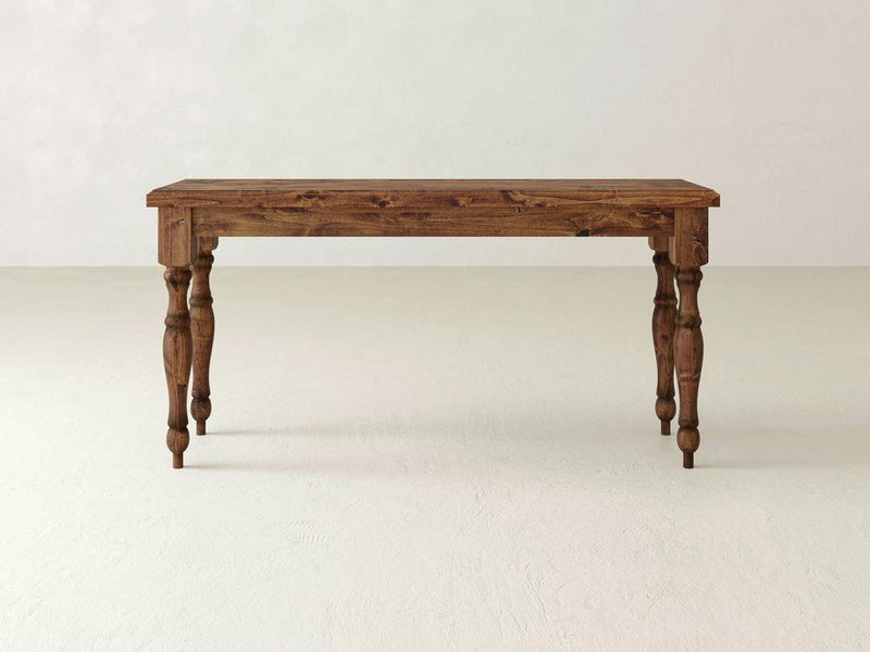 The Abigail Sofa Table - Tuscany by James+James features a rectangular top and four ornately carved legs. It is showcased on a light-colored floor against a plain, light-colored wall. The table's natural, rustic finish beautifully highlights its grain patterns.
