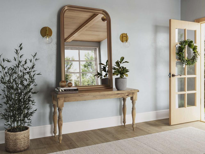 A hallway with an Abigail Sofa Table in Harvest Wheat by James+James, potted plants, a large mirror framed in wood, and wall sconces on either side. An open door with a wreath on it and sunlight streaming in. A woven basket with a tall plant is to the left of the sofa table.