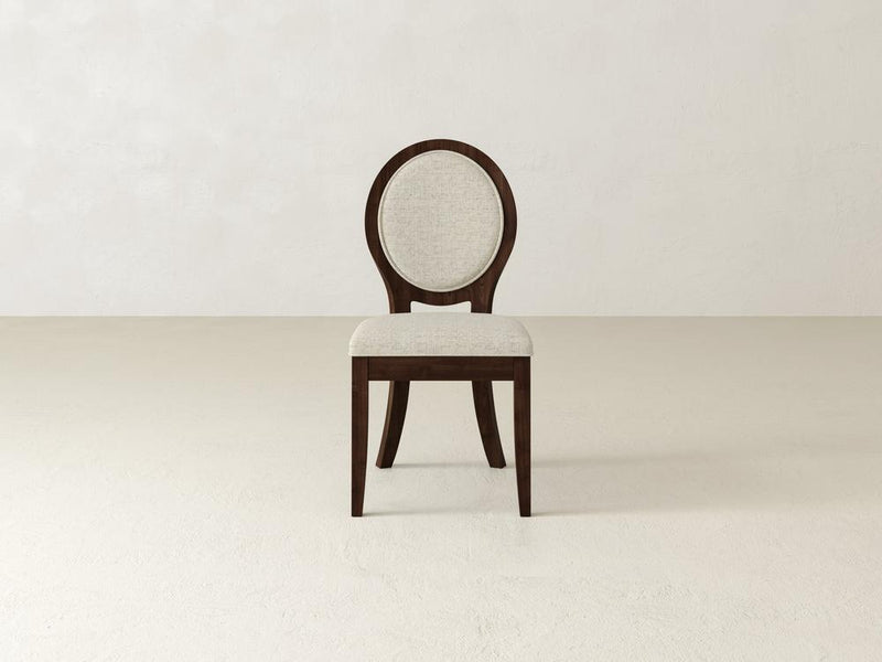 A wooden chair with a round, upholstered backrest and a cushioned seat in light fabric, set against a plain, off-white background. The chair has a dark wood frame and four legs.