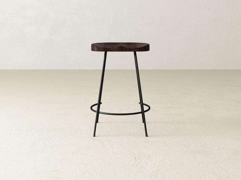 The Bryer Stool by James & James blends rustic charm with its minimalist wooden circular seat and industrial style through its black metal legs, elegantly positioned on a beige concrete floor against a plain light-colored wall.