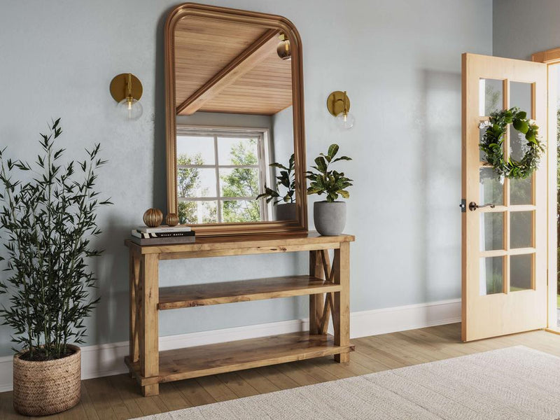 A bright entryway features the Carter X Sofa Table in Harvest Wheat from James+James with a large framed mirror above it. Two plants adorn the table, and a potted plant sits next to it. The open door has a wreath, and the room is filled with natural light, highlighting the light blue walls.