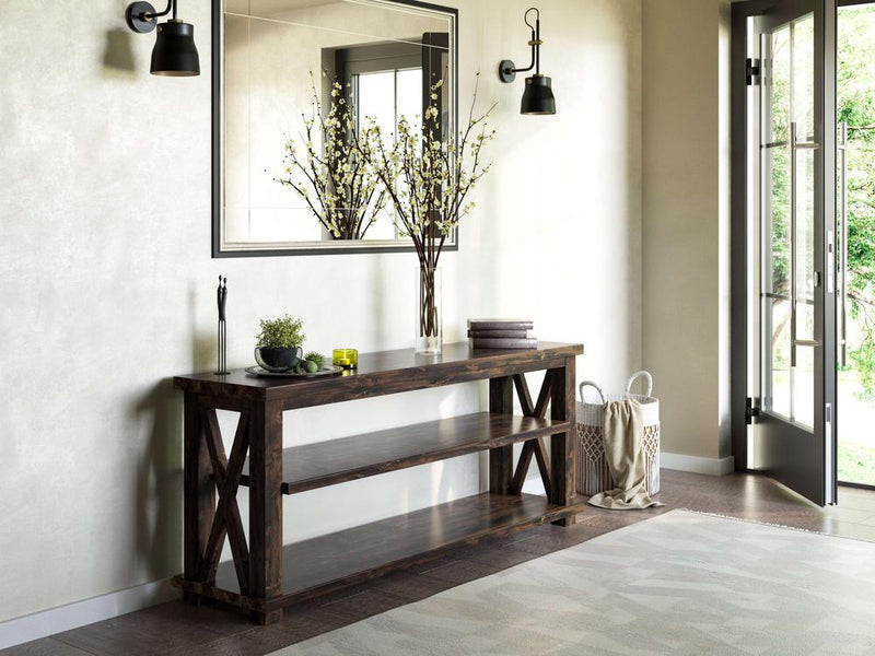 A modern entryway features the Carter X Sofa Table in Tobacco by James+James, with two lower shelves holding a vase with dried branches, a stack of books, and a small tray with decor items. Above the table hangs a large mirror. Black wall sconces flank the mirror, and a door opens to a lush garden view.