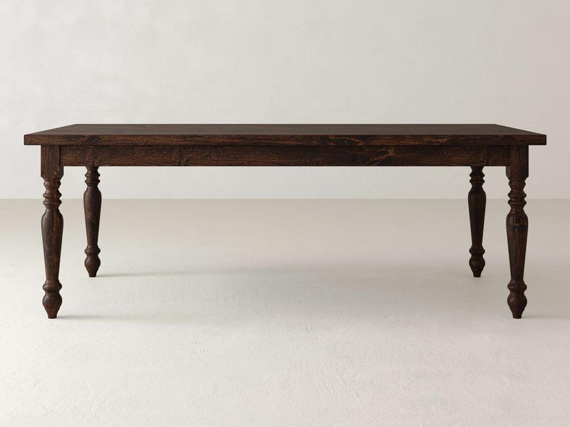 The James+James French Country Dining Table - Tobacco stands on a light-colored floor with its smooth rectangular top and intricately carved legs, set against a plain light wall.