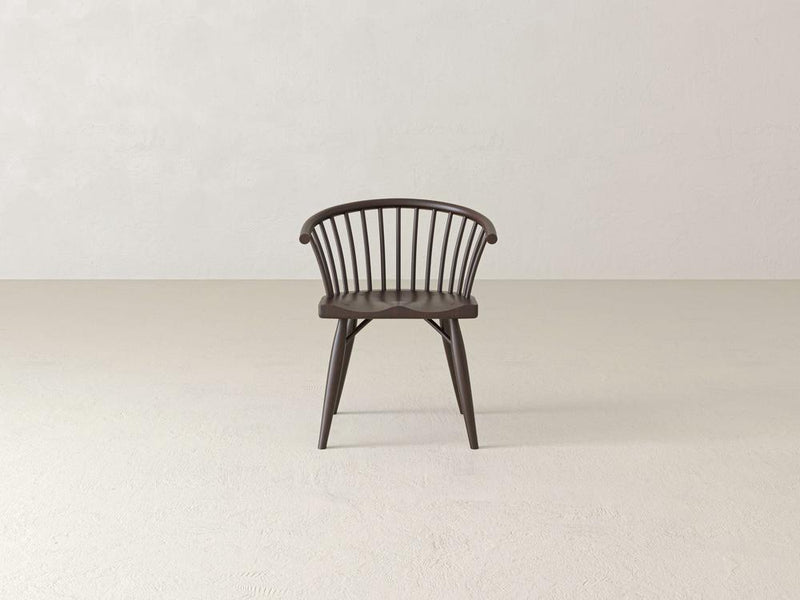 Dark wooden chair with spindle backrest, curved arms, and angled legs placed on a light, textured floor in a minimalist setting.