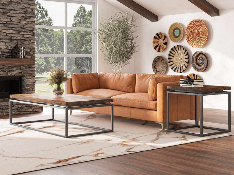A modern living room with a brown leather sectional sofa, two James+James Floating Top End Tables in Tobacco, and a large patterned rug. A display of woven baskets decorates the white wall. There's a tall potted plant by the window, and a stone fireplace in the background.