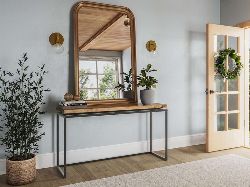 A modern entryway showcases the Floating Top Sofa Table - Harvest Wheat by James+James, with a wooden top and metal legs. The table is adorned with a small plant and a decorative item. A large wooden-framed mirror hangs above it. To the left stands a potted plant, while to the right, a wooden door opens to a bright space.