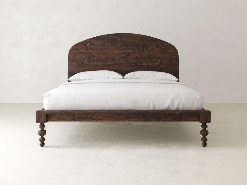 The Olivia Arch Bed - Tobacco from James+James features a wooden frame with an arched headboard and turned legs. It includes two white pillows and a neatly made white bedspread, set against a plain, light-colored wall and floor.