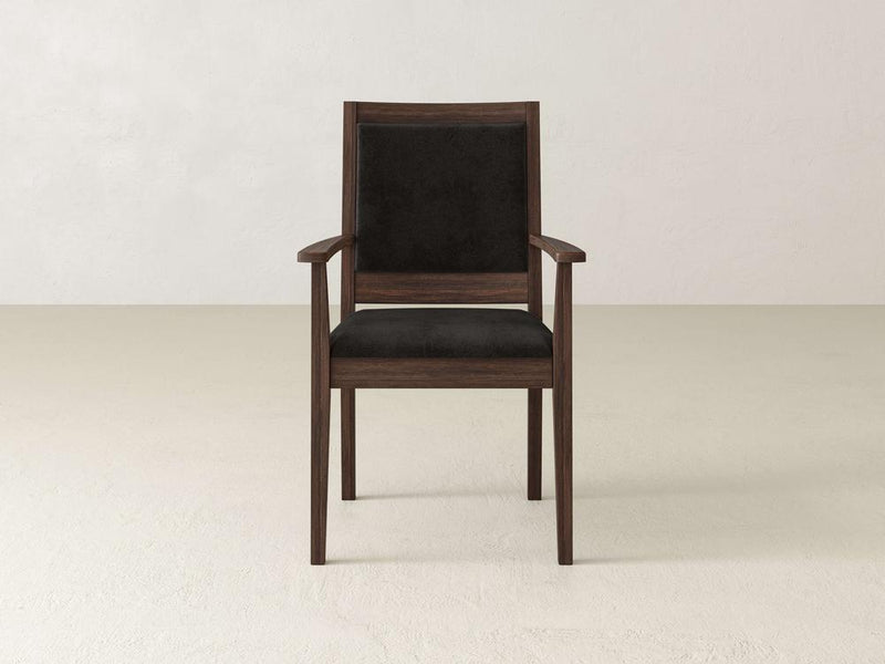 The Gina Arm Dining Chair - Tobacco by James+James, featuring a dark cushioned backrest and seat, rests on a light-colored floor against a plain, light-colored wall. The wooden chair boasts armrests and a minimalist, modern design.