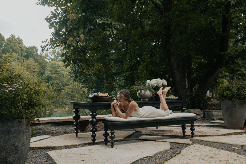 A woman in a white dress lies on her stomach on a James+James Olivia Dining Table - Harvest Wheat, outdoors amid lush green trees and potted plants, gazing thoughtfully ahead in a tranquil garden setting.