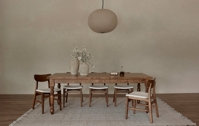 A minimalist dining room features the James+James French Country Dining Table - Tuscany, four wooden chairs with white cushions, a beige rug, white-flowered vases as decor, and a large round paper pendant light for rustic charm.