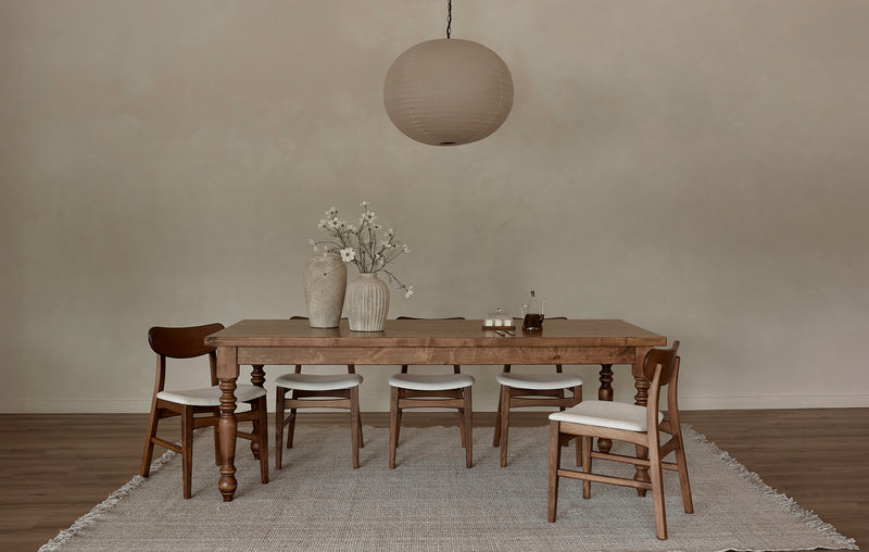 A minimalist dining room features the James+James French Country Dining Table - Tuscany, four wooden chairs with white cushions, a beige rug, white-flowered vases as decor, and a large round paper pendant light for rustic charm.
