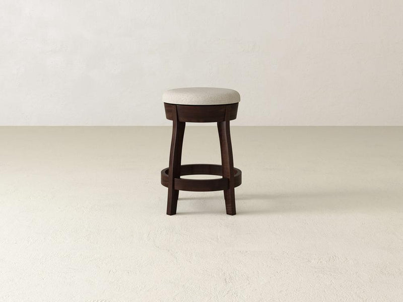 The James & James Jasper Swivel Stool has a wooden base, beige upholstered cushion, circular footrest, and four sturdy legs. It's elegantly displayed on a light surface against a neutral wall.