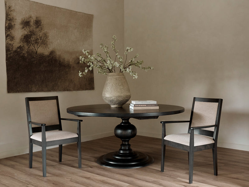 The Josephine Round Dining Table - Tuscany by James+James sits between two beige upholstered chairs, topped with a large rustic vase of branches and two books. A sepia landscape artwork hangs above the wood flooring.