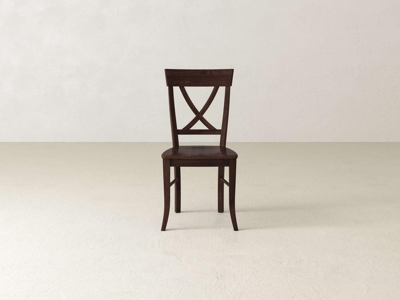 A wooden chair with a dark finish stands on a light-colored floor against an off-white wall. The chair features a simple design with an X-shaped backrest and four straight legs.