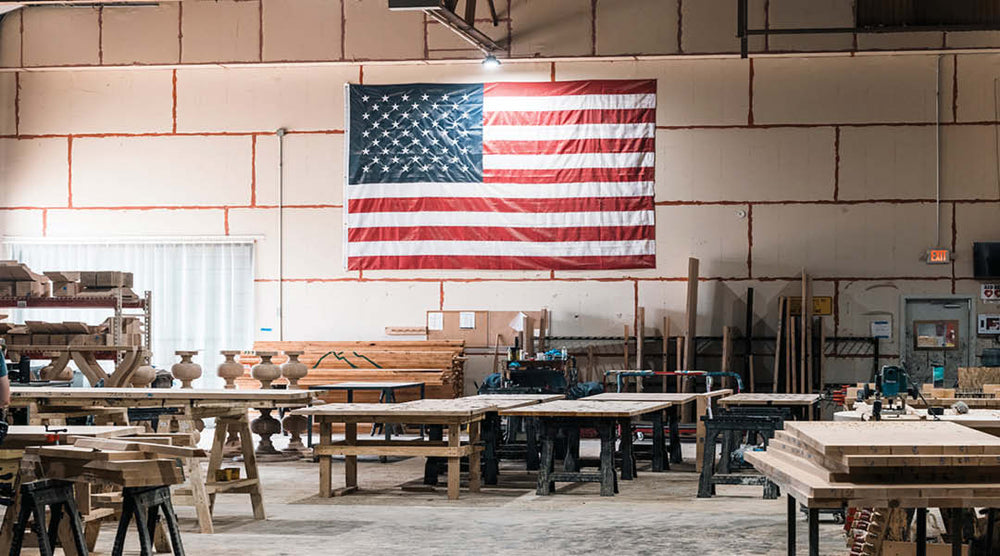 A large American flag hangs on the wall of a spacious woodworking shop. The room is filled with workbenches and tools. Soft lighting illuminates the area, creating a warm and industrious atmosphere.