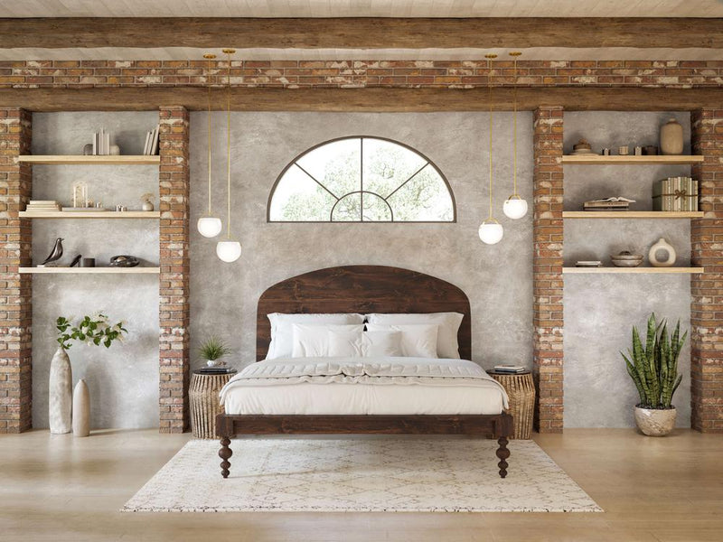 A cozy bedroom with a rustic vibe features the Olivia Arch Bed in Tobacco by James+James and white bedding. Brick walls with built-in shelves hold decor. An arched window, pendant lights, a plant, and a soft rug complete the scene.