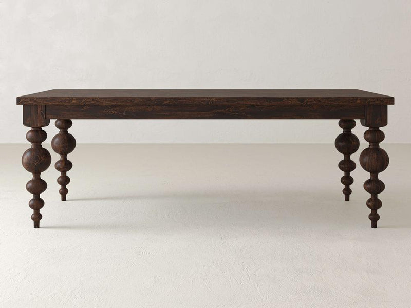 The Olivia Dining Table - Tobacco by James+James is a rectangular wooden table featuring intricately carved, spherical legs in a dark finish. It is set against a light-colored wall and rests on a smooth, light-toned floor.