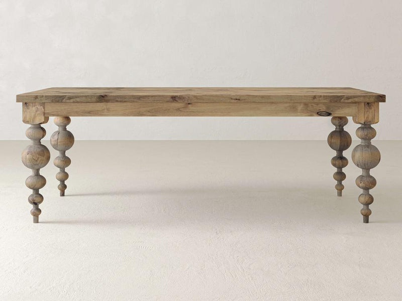 The Olivia Dining Table - Harvest Wheat by James+James features intricately turned legs and stands on a plain, light-colored floor. The tabletop boasts a natural finish, beautifully highlighting the wood grain. In the background is a smooth, neutral-colored wall.