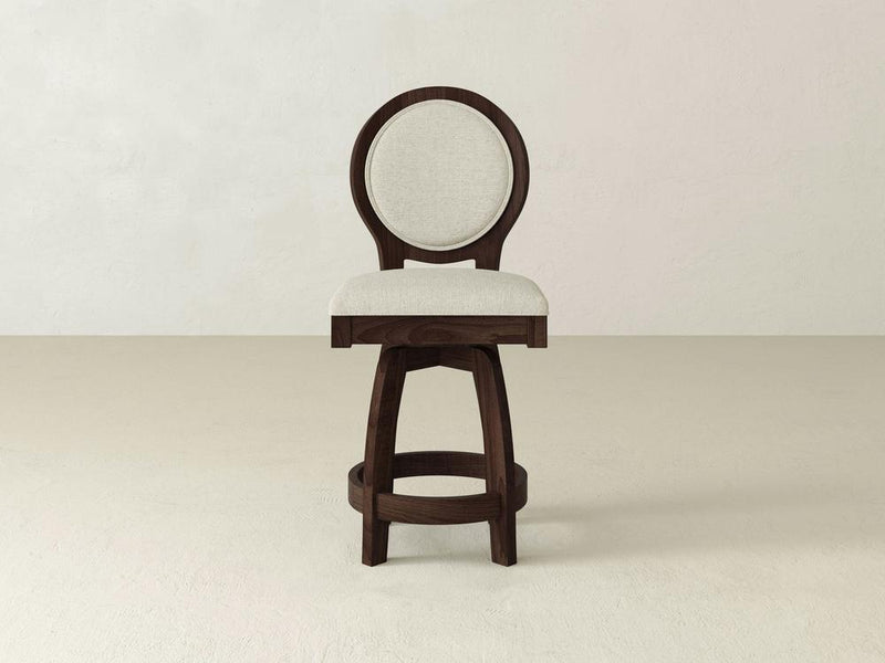 The James & James Alice Swivel Stool features a circular upholstered backrest and seat against an off-white backdrop. With a maple wood frame and round base support, it enhances stability and complements kitchen counters with its classic design.