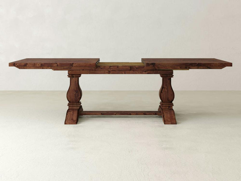 The Heirloom Expandable Dining Table - Tuscany by James+James is a wooden dining table with a rustic design. It features a rectangular top that is divided in the middle, suggesting the presence of a removable leaf extension. The base consists of two ornate legs connected by a horizontal support, set against an off-white wall background.