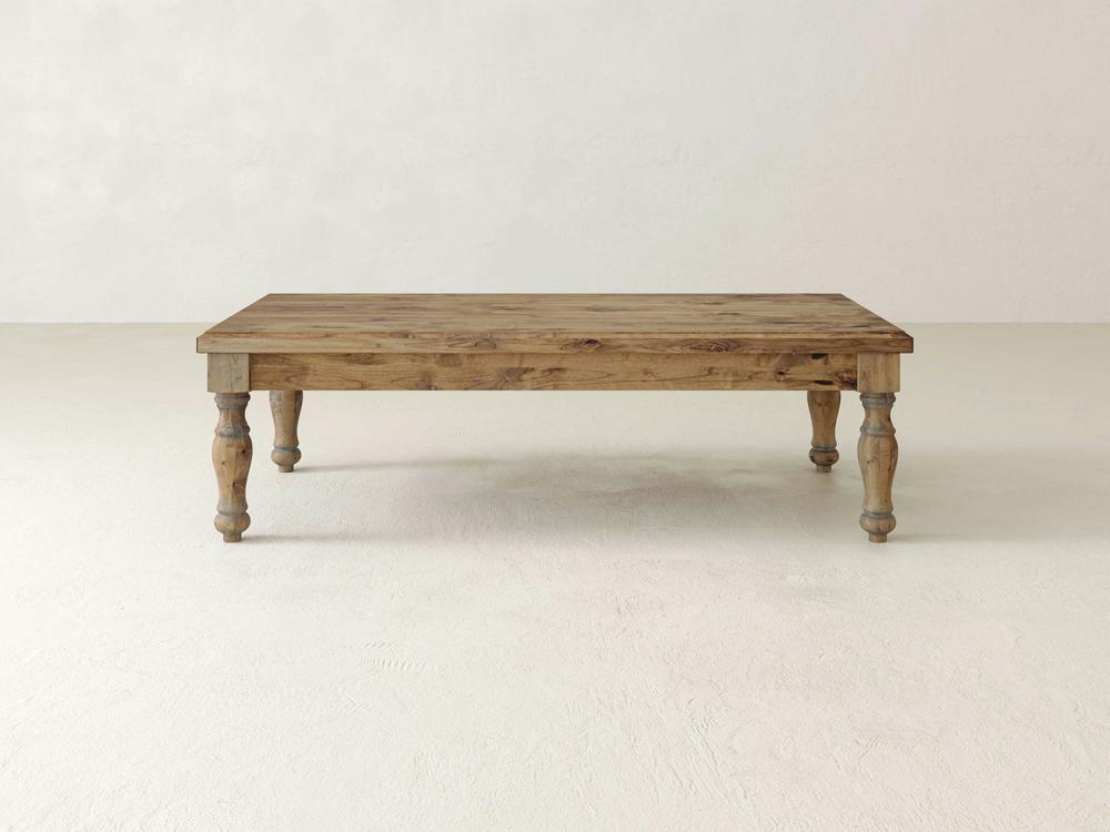 The **Abigail Coffee Table - Harvest Wheat** by **James+James** features a rectangular wooden design with a rustic finish. It stands on four intricately carved legs and boasts a smooth surface that showcases the visible wood grain. The table is placed on an off-white floor against a plain, light-colored background.