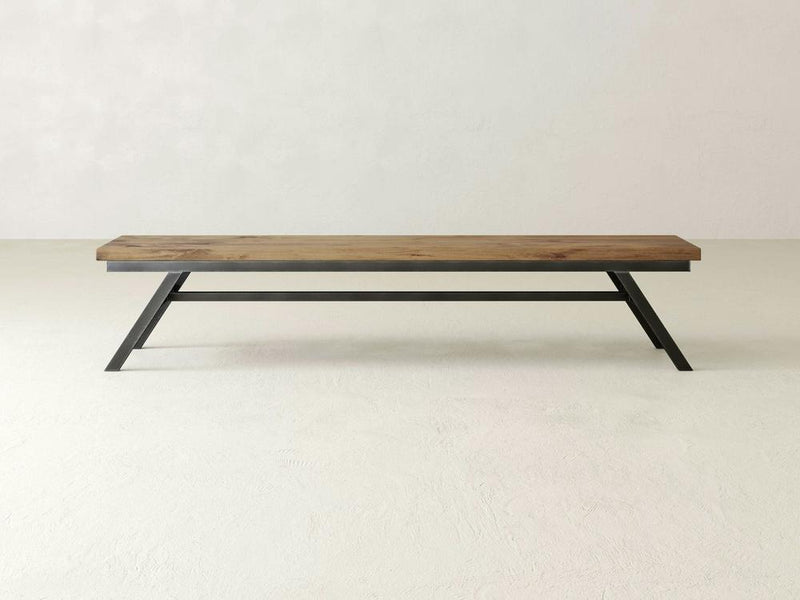 The Fulton Bench - Harvest Wheat by James+James is set against a plain, off-white background. This minimalist wooden bench features a rustic, natural finish and has a single plank seat supported by black, angular metallic legs. The design is simple and modern, emphasizing clean lines and functionality.