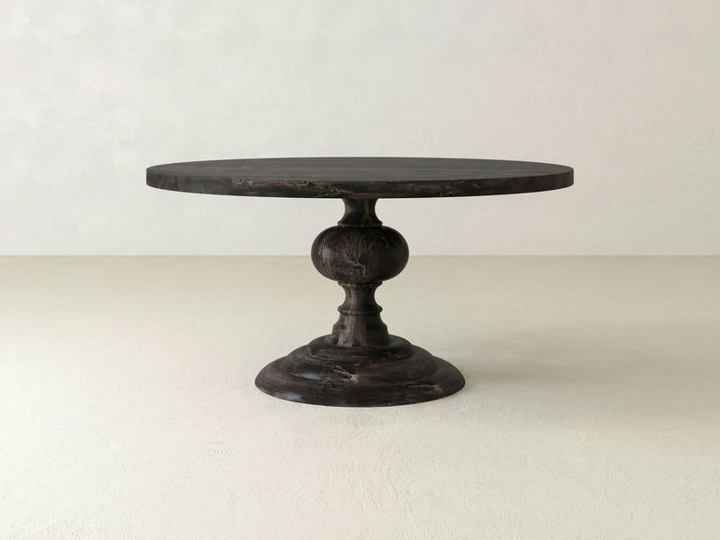 The Josephine Round Dining Table - Charred Ember from James+James is a round, dark-colored wooden table featuring a single pedestal base with an ornate design and a wide, sturdy bottom. It boasts a classic and elegant look, perfect for dining or decorative use against the plain white background.