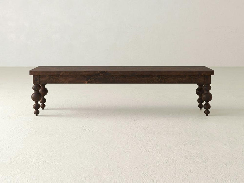 The Olivia Bench - Tobacco by James+James, a long, rectangular wooden bench with a dark brown finish, is centered against a plain, light gray background. The bench features four intricately carved, baluster-style legs that add a touch of elegance and vintage charm to its simple design.