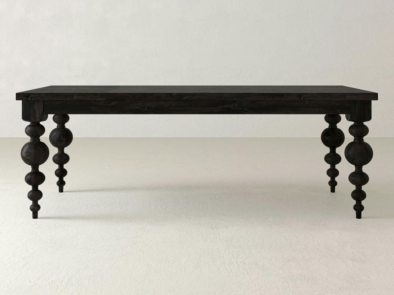 The Olivia Dining Table - Charred Ember by James+James is a dark wooden table with intricately carved legs featuring both spherical and cylindrical shapes. Its rectangular tabletop stands out against a plain, light-colored wall, presenting a minimalist yet elegant design.
